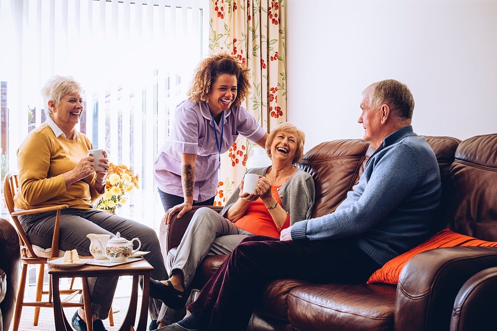 Three seniors enjoying tea and cake in the care home. There is a caregiver laughing with them.