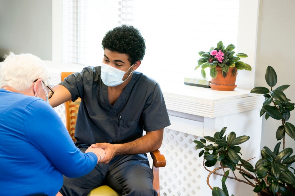 Portrait of a smling African-American male nursing employee wearing gray scrubs and face mask interacts with a senior aged patient during the Covid-19 pandemic, Midwest, USA